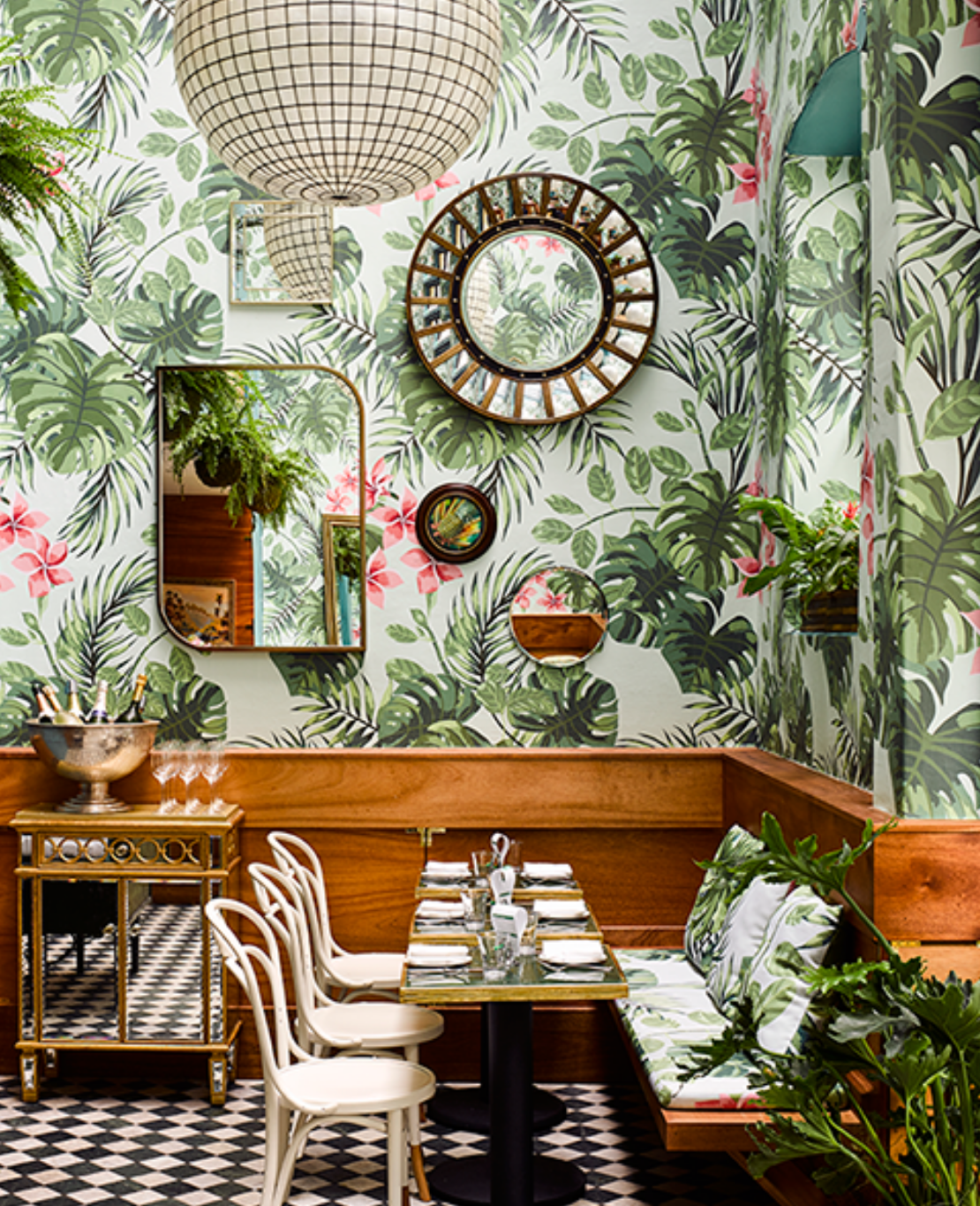 2ND 1st DRAFT LEOS WEBSITE- 4.4.2024 (1) A vibrant restaurant corner with tropical wallpaper, wood paneling, a checkered floor, and stylish mirrors, featuring a set dining table and hanging plants.